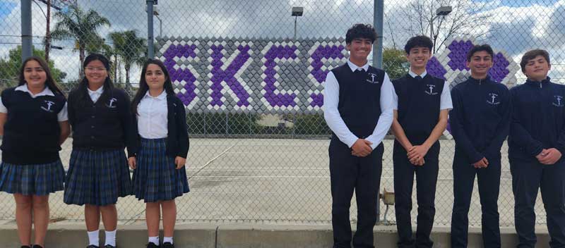 7 students in front of a fence sign that reads SKCS 